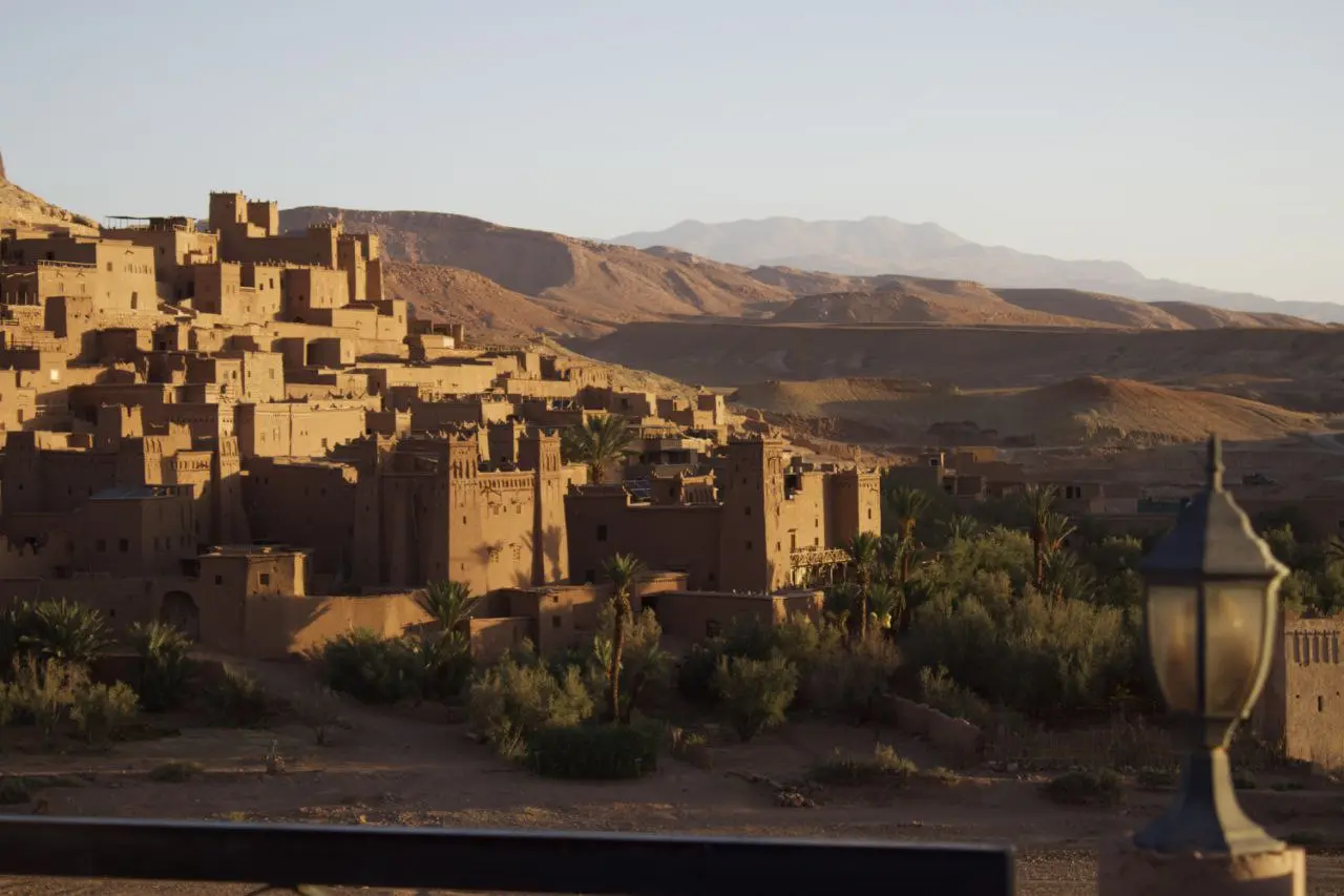 The ancient mud-brick ksar of Ait Benhaddou in Morocco at sunset, surrounded by palm trees and desert hills.