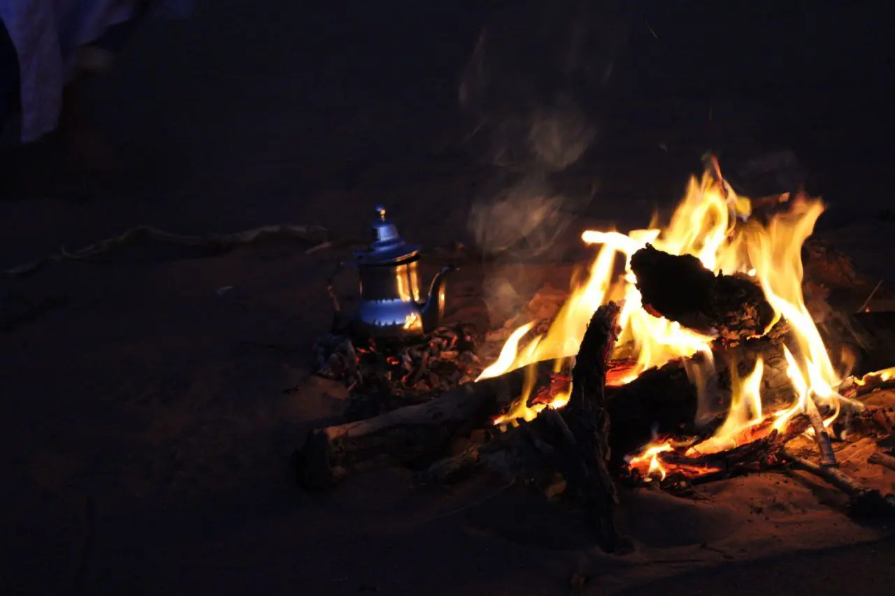 A bright campfire and a silver teapot on embers in the desert sand at night.