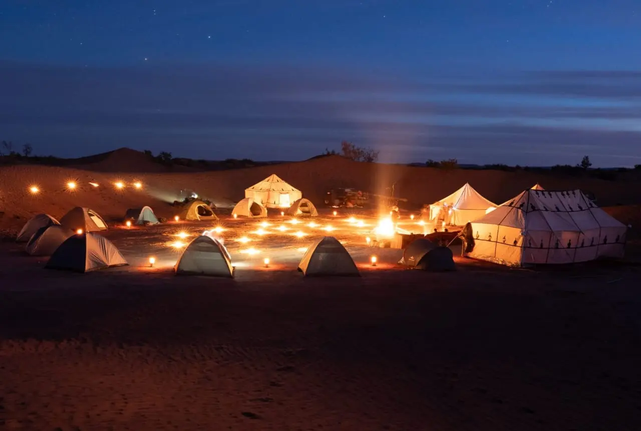 Night view of a desert camp with various tents and a central campfire, illuminated by numerous lanterns under a starry sky.
