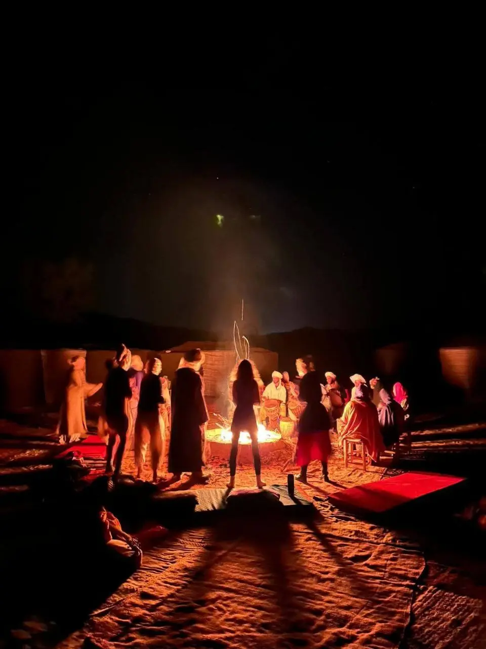 A group of people gathered around a large campfire in a desert at night, silhouetted by the warm orange glow.