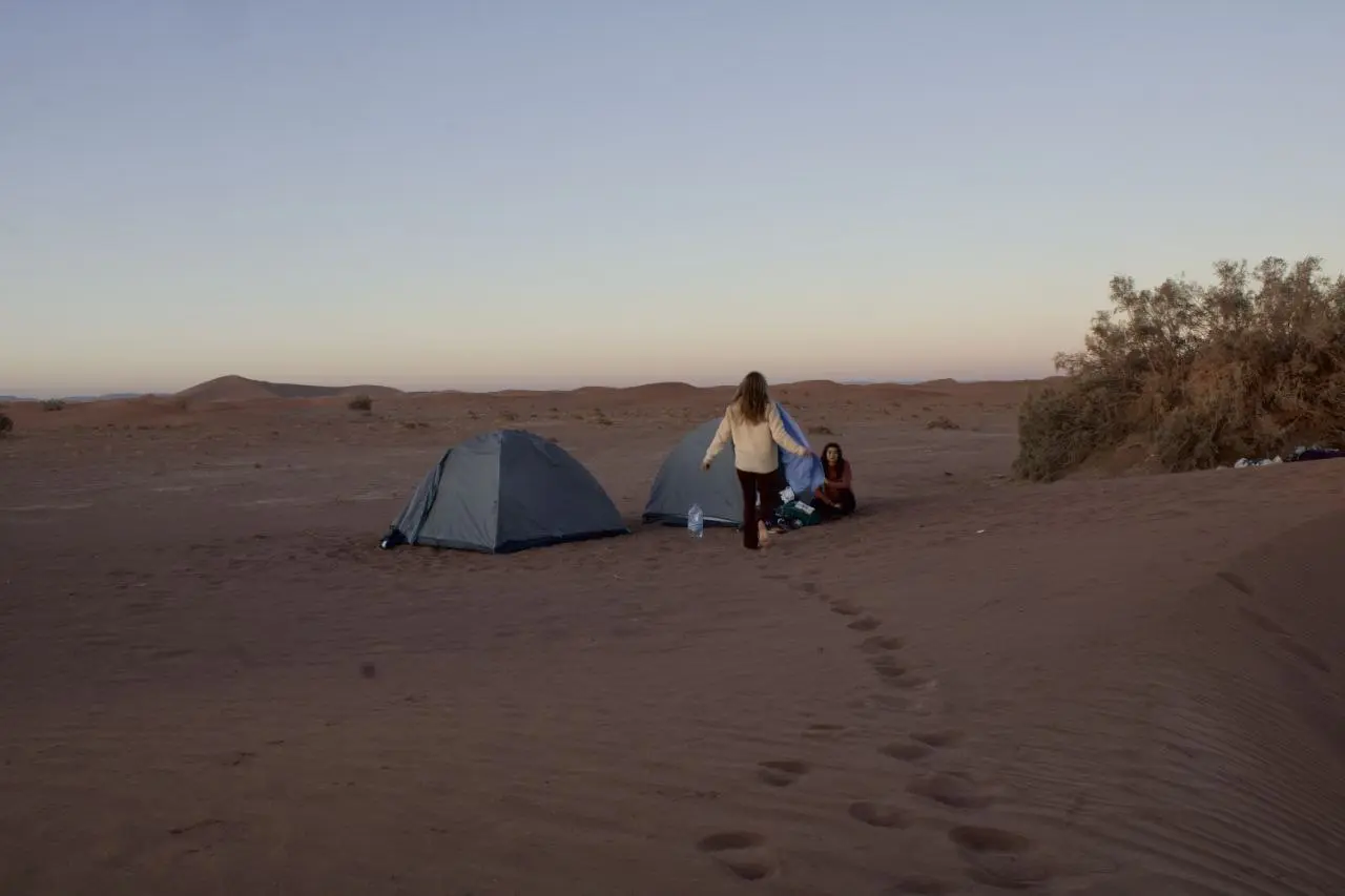Two people camping with grey tents in a vast desert landscape at dusk with footprints in the sand.
