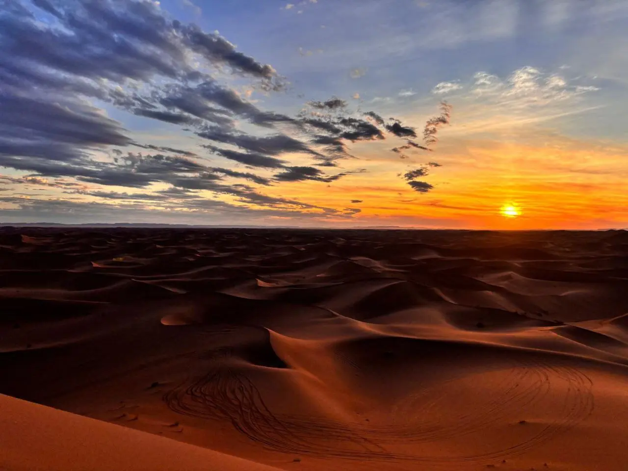 Dramatic desert sunset with a vivid orange and yellow sky, dark blue clouds, and endless reddish-brown sand dunes.