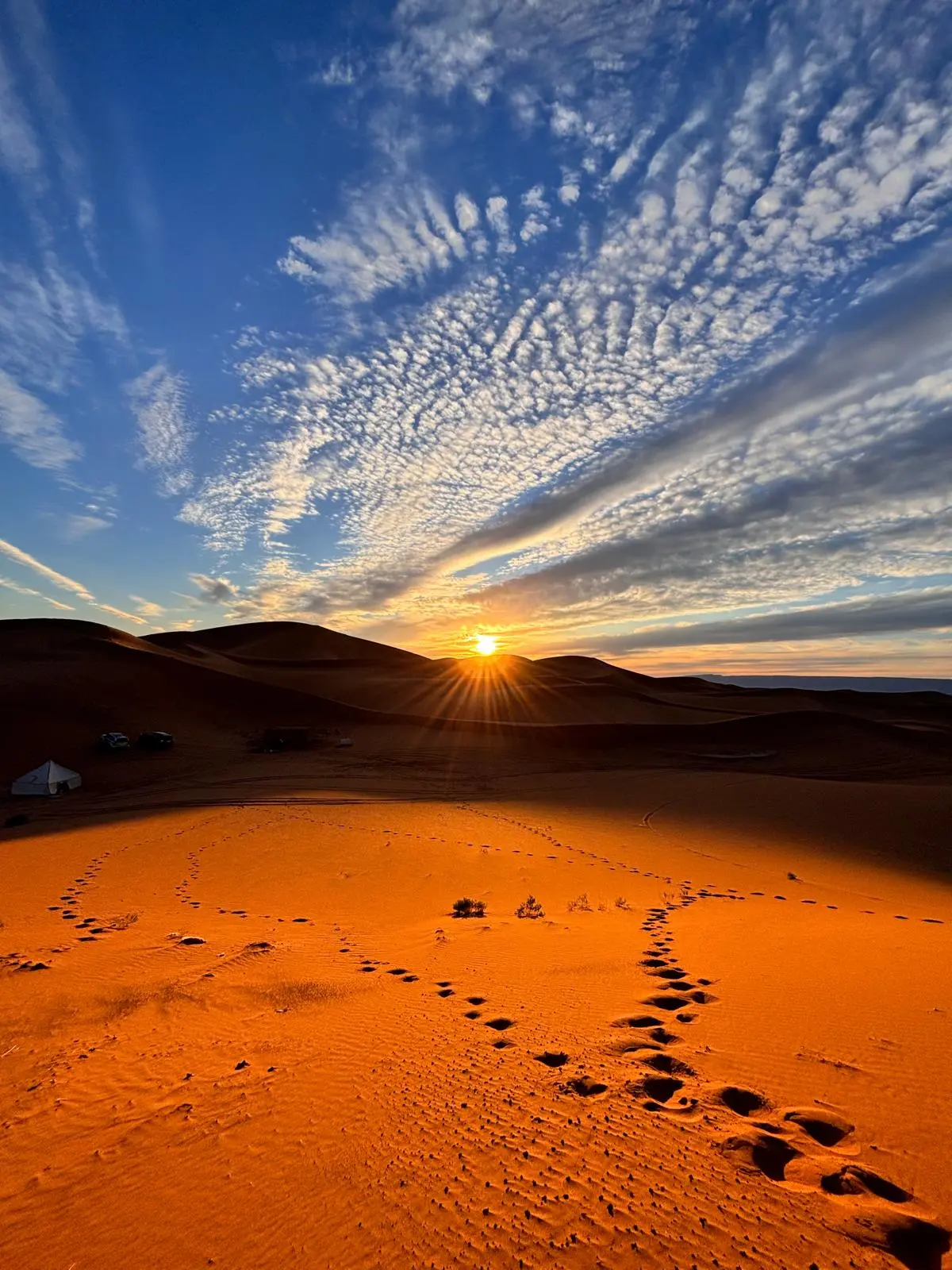 Sunrise or sunset over a desert camp with orange-red dunes, starburst sun, and rippled clouds.