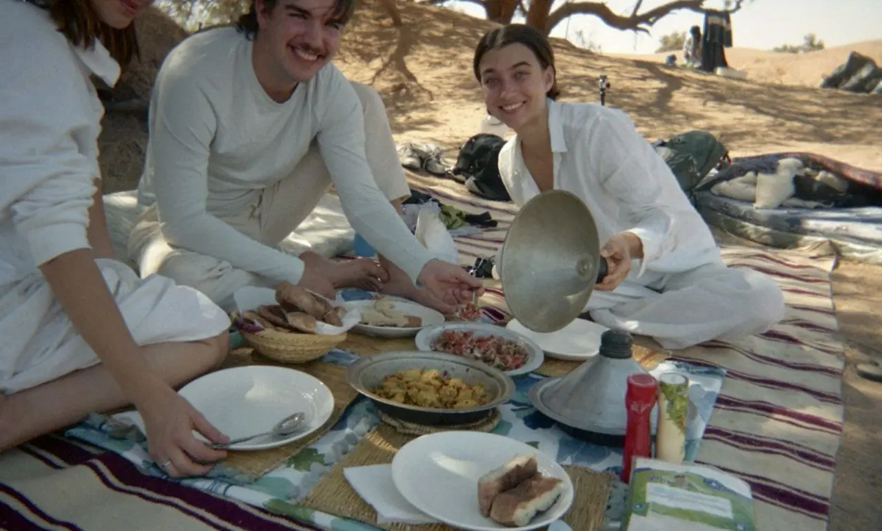Three friends in white clothing share a traditional Moroccan meal on a rug in the desert.