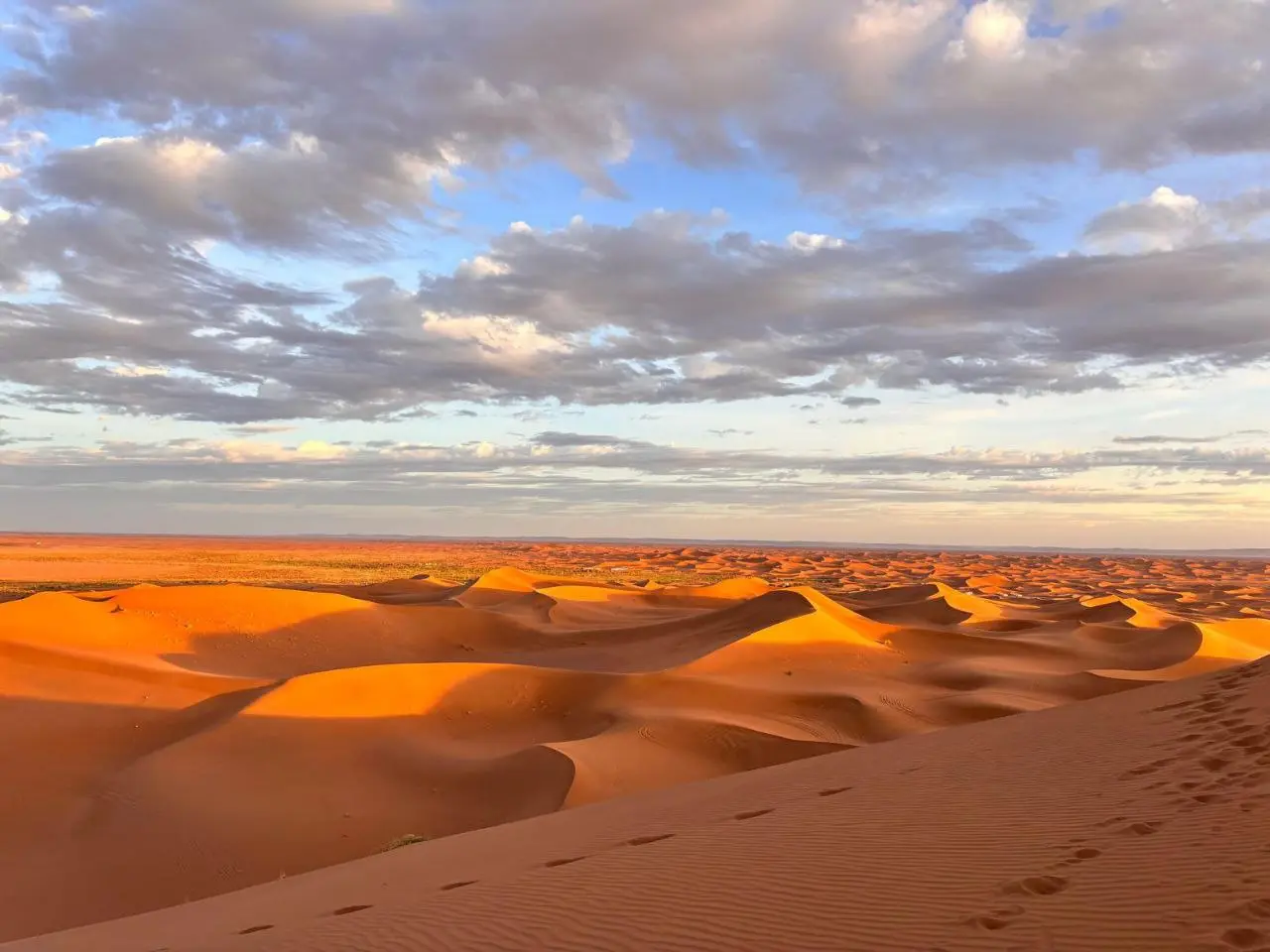 Golden sand dunes under a cloudy sky at sunset with footprints in the foreground.