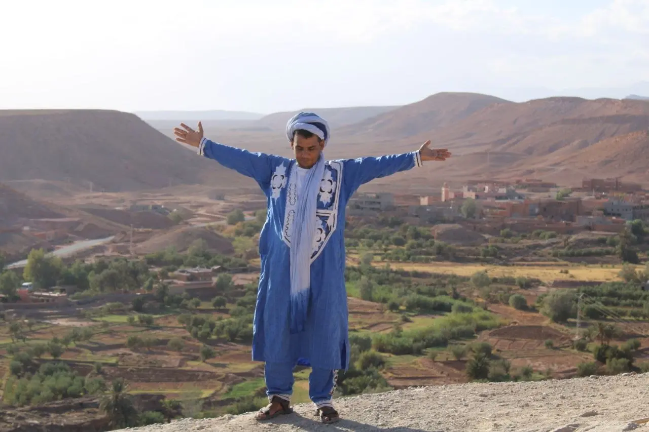 Man in blue traditional clothing with arms outstretched overlooking a vast desert valley and mountains.