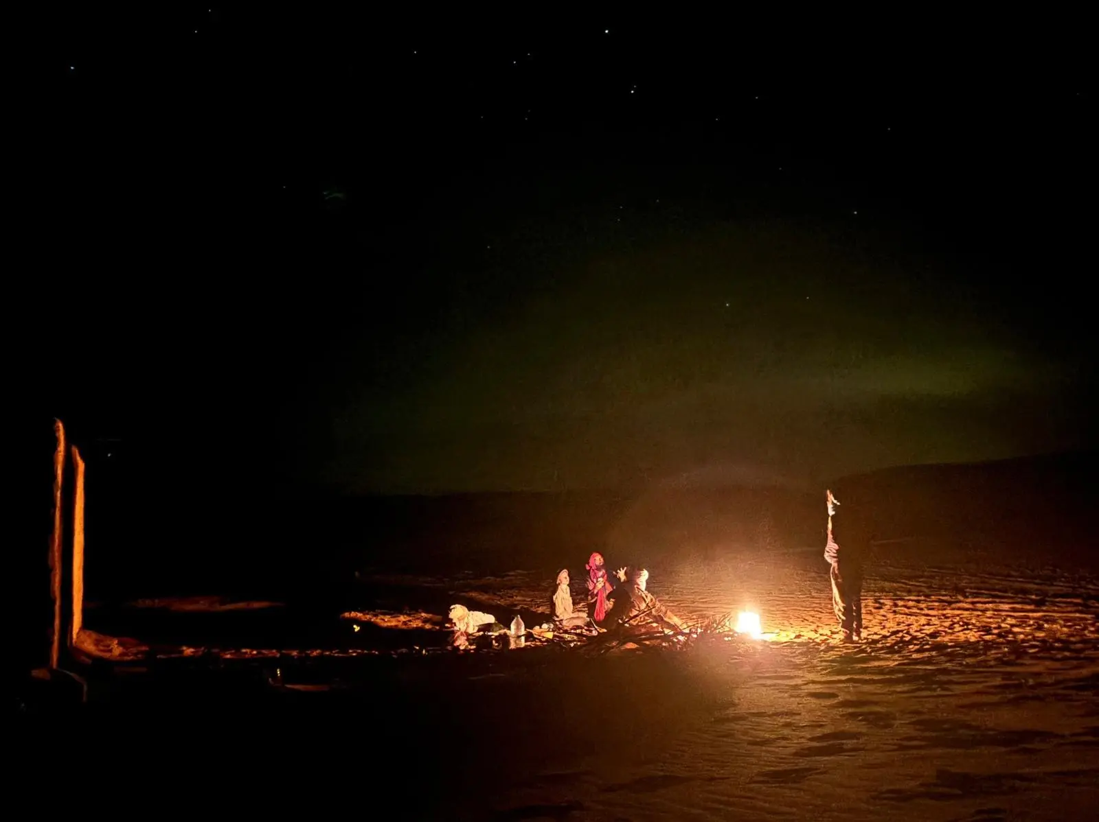 Night sky with soft green aurora above people and dog around a bright campfire in desert.