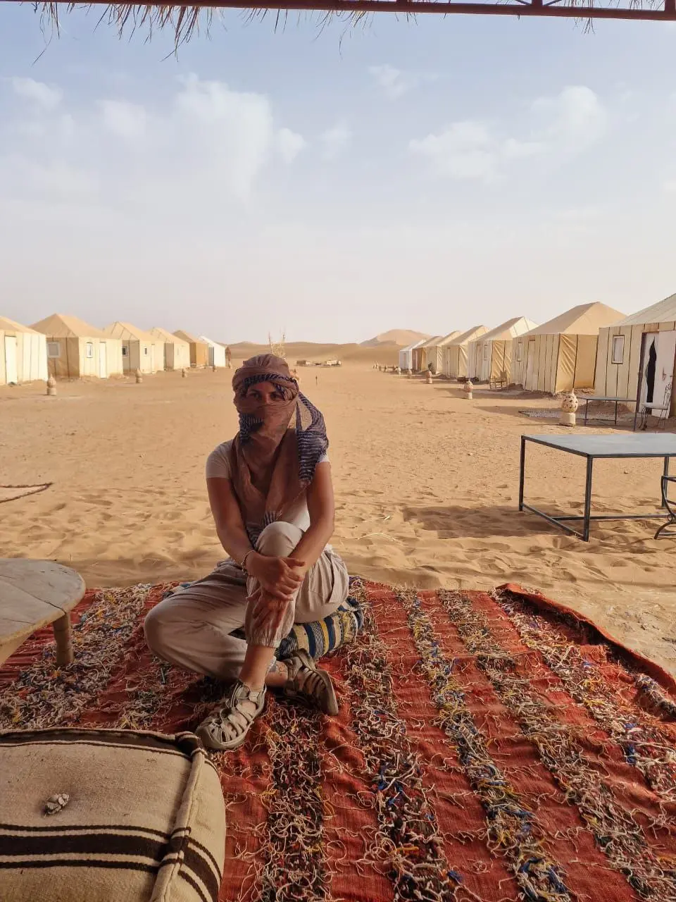 Person with a headscarf sitting on a rug at a desert camp with rows of tents in the background.