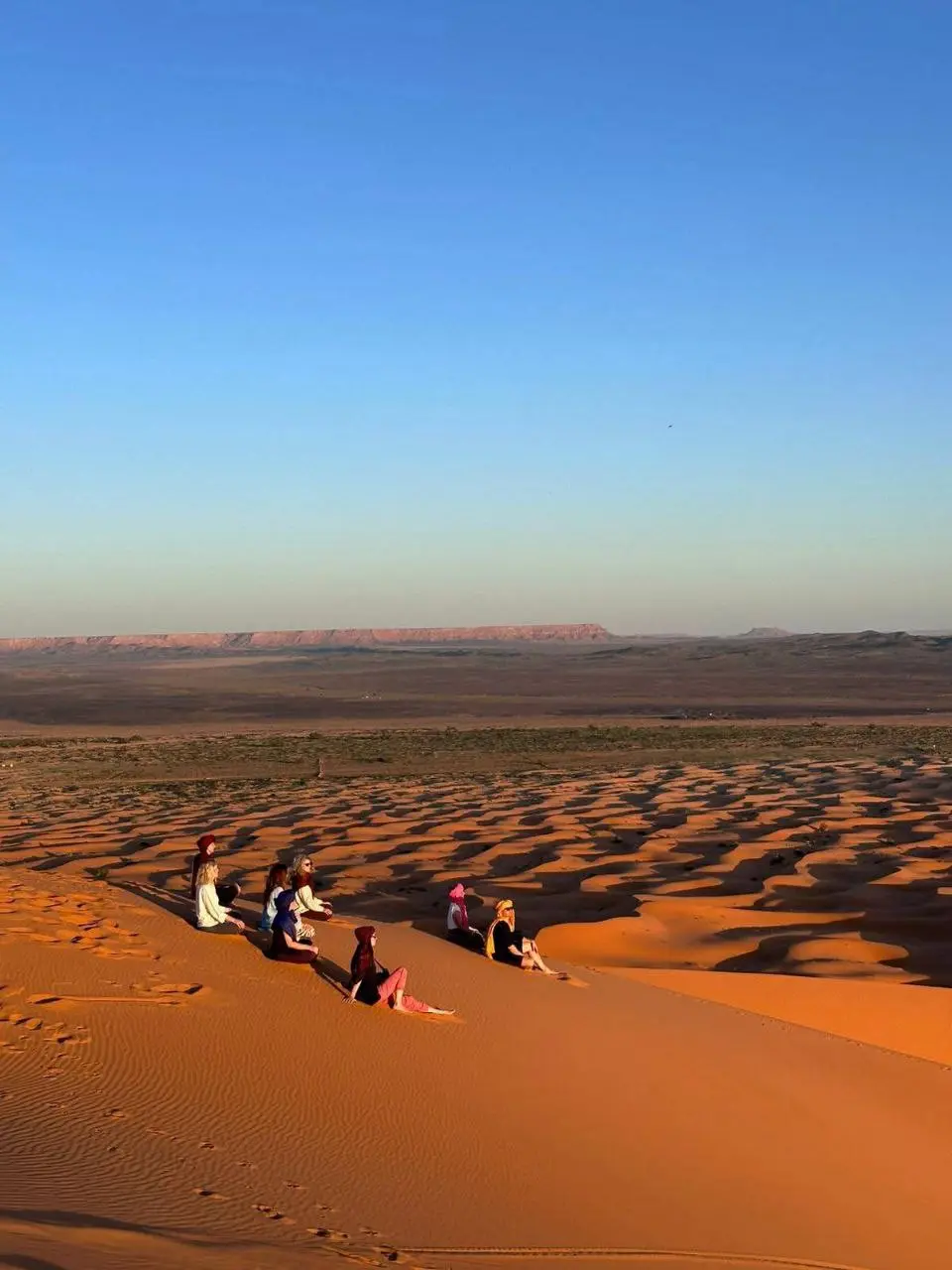 A group of people sit on a large orange sand dune, looking out over a vast desert landscape under a clear blue sky.