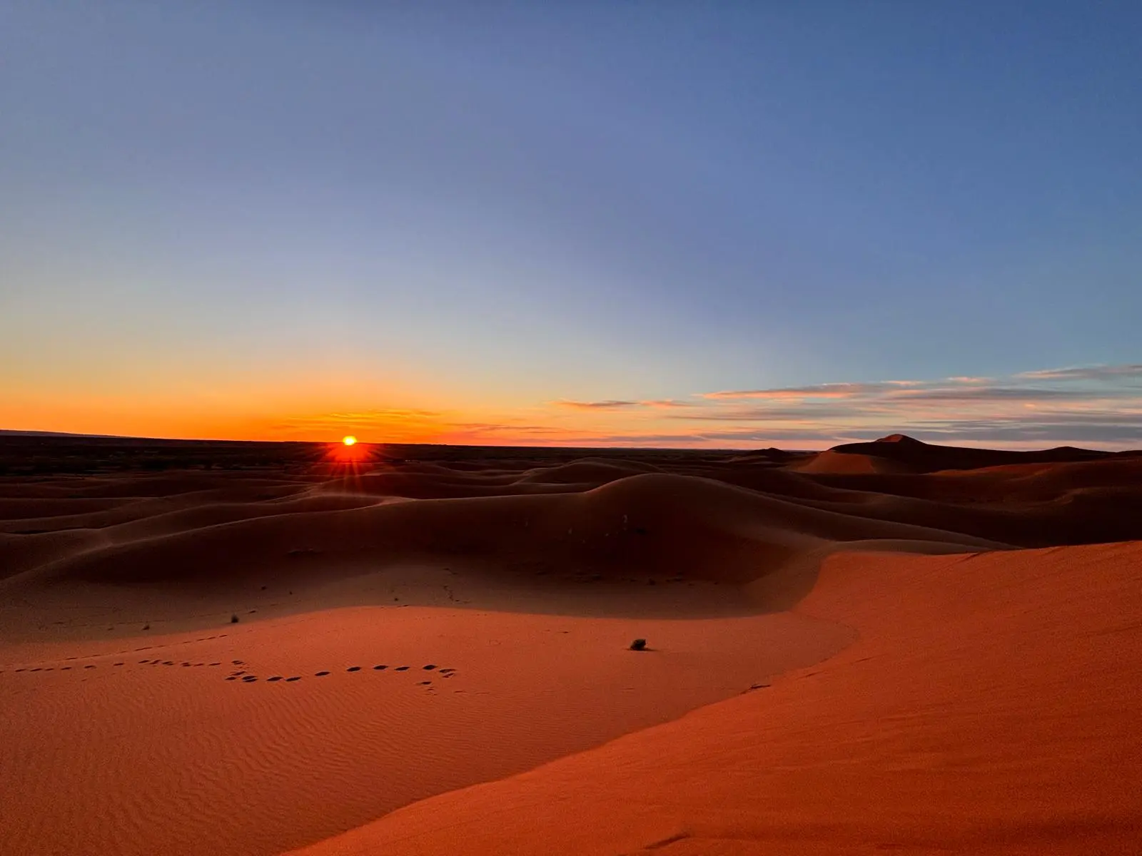 Striking desert landscape at sunset with vibrant orange sun on the horizon above undulating sand dunes.