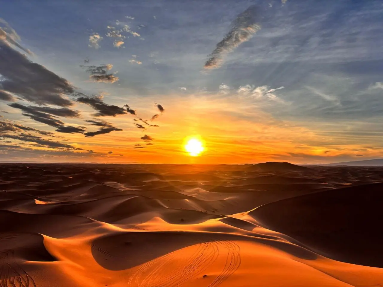 A vibrant golden sunset over rolling sand dunes in a vast desert landscape under a wispy sky.